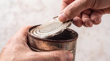 Dos manos sostienen y abren una lata de metal oxidada, mostrando la tapa levantada y el borde interior del recipiente.
