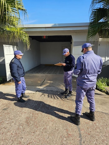 Cuatro hombres con uniforme policial azul y violeta en un exterior. Uno sostiene un portapapeles. Se encuentran frente a un garaje abierto