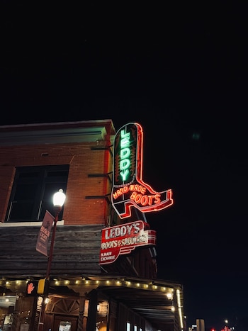 Rótulo de neón de bota cowboy en fachada de edificio, con textos "Leddy's Hand Made Boots" y "Boots & Saddle". Farola con bandera "Busch Light Rodeo"