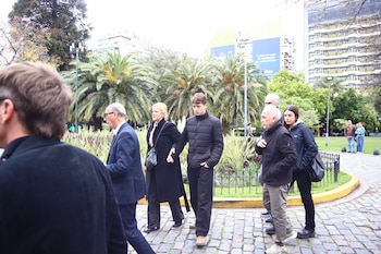 Varias personas vestidas con abrigos oscuros caminan por un camino de adoquines cerca de un parque con palmeras