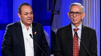 Composición de dos hombres en trajes oscuros. El de la izquierda con camisa blanca y pin rojo; el de la derecha con corbata roja y gafas. Ambos ante micrófonos.