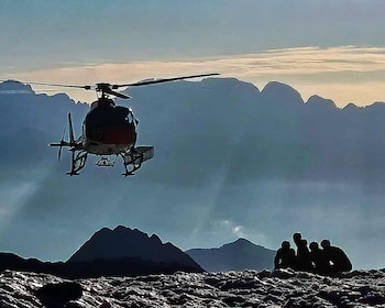 Un helicóptero en vuelo frente a siluetas de montañas y cuatro personas sentadas sobre un terreno rocoso