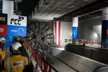 Cabezal de la tuneladora "Delia" emergiendo en una estación de metro en construcción. Obreros con cascos y chalecos amarillos observan. Pilares, vías y una bandera peruana