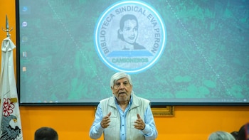 Hugo Moyano, hombre mayor de pelo blanco y chaleco beige, habla gesticulando. Detrás, una pantalla muestra el logo de la Biblioteca Sindical Eva Perón y una bandera