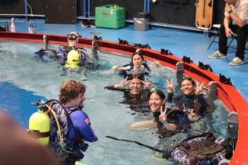 Personas en una piscina azul. Algunos con equipo de buceo, incluyendo tanques amarillos. Jóvenes sonríen y hacen el signo de la paz. Un instructor y otro hombre observando