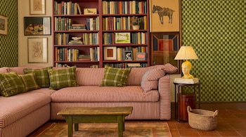 Sofá seccional rojo con rayas, cojines verdes de cuadros, mesa de centro de madera, librería roja con libros, cuadros, lámpara de mesa, cesta de mimbre.