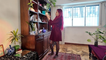 Mujer con cabello magenta y vestido granate observa un objeto en una estantería de madera oscura con libros y plantas. Hay una ventana al fondo
