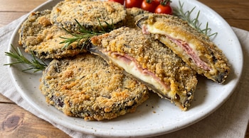 Plato blanco con filetes de berenjena empanados, algunos rellenos de jamón y queso, ramitas de romero y tomates cherry. Se observa un mantel y una mesa de madera.