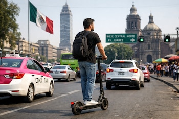 Hombre de espaldas con mochila negra en monopatin eléctrico en avenida de Ciudad de México. Se ven coches, bandera mexicana, Torre Latinoamericana y edificios históricos.