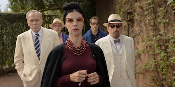Una mujer con turbante negro, gafas, collar rojo, jersey granate y cárdigan oscuro, con cuatro hombres vestidos con trajes claros y sombreros
