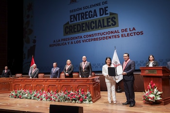 Una mujer en traje blanco recibe un documento de un hombre en un escenario con la bandera de Perú. Detrás, una pantalla muestra "Entrega de Credenciales"