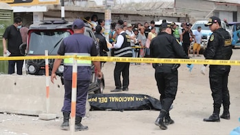 La policía y personal forense procesan la escena de un crimen. Un cuerpo en bolsa negra yace en el suelo cerca de un mototaxi. Hay cinta policial amarilla y curiosos