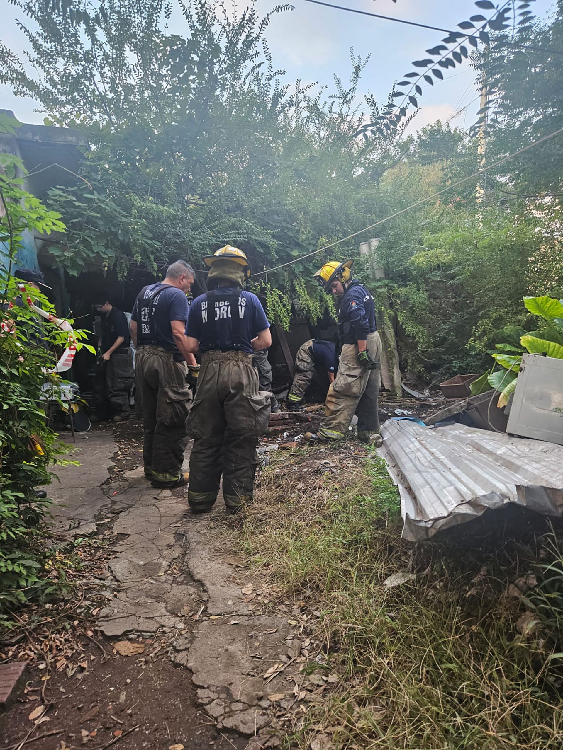 Personal de emergencia y peritos trabajan en un área exterior con vegetación densa y escombros, como parte de la investigación del crimen del rapero Mike Dee