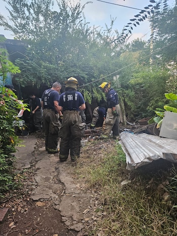 Varias personas con uniformes de trabajo y cascos inspeccionan un área exterior cubierta de vegetación, tierra y restos de material