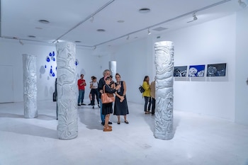 Sala de exposición de arte moderna con paredes blancas, varias esculturas cilíndricas blancas y obras abstractas azules y grises. Hay visitantes admirando