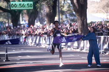Un atleta cruza una cinta morada de llegada que sostiene una voluntaria bajo un cronómetro digital iluminado en verde