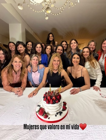 Un grupo de mujeres posa detrás de una mesa blanca con un pastel decorado con fresas y velas bajo una lámpara colgante