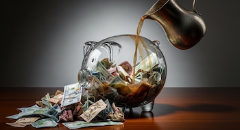 Una jarra vierte líquido oscuro en una alcancía de cristal llena de billetes de varias monedas, con más dinero desparramado en una mesa de madera.