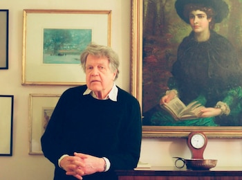 Merlin Holland, un hombre mayor con cabello gris y suéter oscuro, posa en interiores frente a una pintura clásica de una mujer y otros cuadros