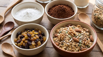 Tazones con polvo de hornear, cacao en polvo, pasas de uva rubias y negras, y granola con avena, frutos secos y semillas. Cucharas de madera.