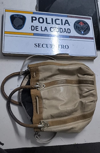 Primer plano de una cartera de mano beige y marrón sobre una superficie blanca, con un cartel de la Policía de la Ciudad que dice 'SECUESTRO' detrás