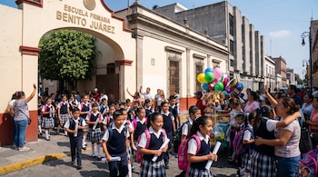 Estudiantes con uniforme escolar salen por el arco de la Escuela Primaria Benito Juárez junto a adultos y un vendedor de globos.