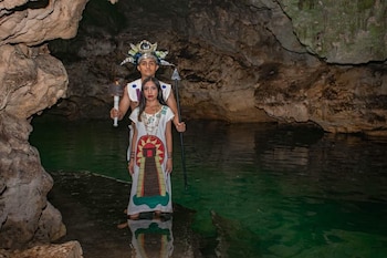 Un hombre y una mujer con vestimenta ceremonial posan en una cueva con agua. El hombre sostiene una antorcha encendida. La mujer lleva un vestido estampado
