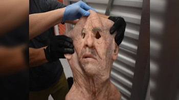 Dos personas con guantes de goma sostienen una máscara flexible con facciones de anciano frente a una persiana metálica