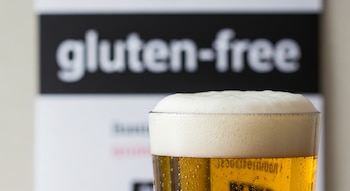 Primer plano de un vaso de cerveza con espuma y burbujas. Al fondo, una etiqueta con las palabras "gluten-free" en blanco sobre negro, ligeramente desenfocada.