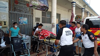 Varios bomberos mueven una camilla con una persona cubierta por una cobija frente a un hospital y junto a una ambulancia