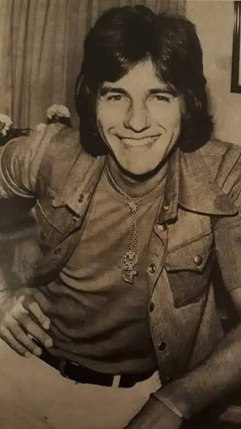 Fotografía en blanco y negro de un joven Sergio Denis, con cabello oscuro y largo, chaqueta de jean, camiseta clara y un collar con colgante grande