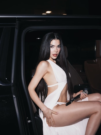 Una mujer de cabello largo oscuro con un vestido blanco sostiene un micrófono sentada dentro de un vehículo negro
