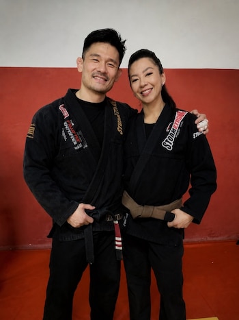 Un hombre y una mujer vestidos con atuendos negros de artes marciales posan abrazados ante un fondo rojo y blanco