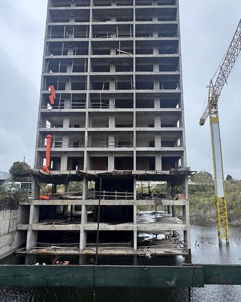 Vista aérea de un edificio de hormigón de muchos pisos sin terminar, con andamios visibles en la parte superior, rodeado de árboles y edificios bajos