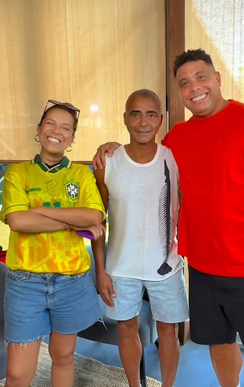 Tres personas posan sonriendo en un interior: una mujer con camiseta amarilla, Romário con camiseta blanca y Ronaldo con camiseta roja