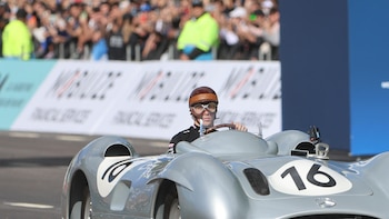 Un hombre con casco y gafas de época conduce un coche de carreras clásico plateado con el número 16 en el lateral, con una multitud borrosa al fondo