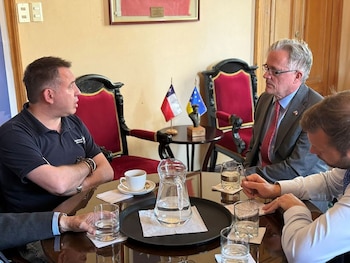 Cuatro hombres sentados a una mesa redonda con vasos de agua, una jarra, una taza de café y dos pequeñas banderas al fondo