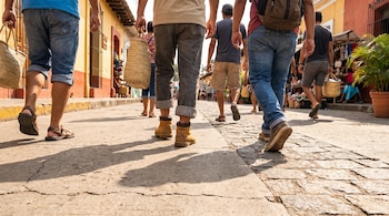 Vista de las piernas de varias personas caminando por una calle de piedra y tierra con edificios de colores y un puesto de mercado al fondo, llevando sandalias y botas.