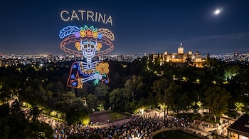 Figura luminosa de una catrina formada por drones en el cielo nocturno sobre un parque arbolado, un castillo iluminado y una multitud.