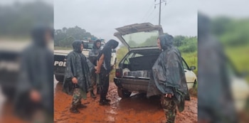 Agentes con ponchos de lluvia detienen a un hombre esposado en camino de tierra rojiza bajo la lluvia. Auto blanco con maletero abierto y paquetes es visible