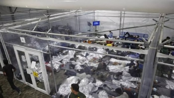 Vista aérea de un amplio centro de detención temporal con muchas personas acostadas en el suelo cubiertas con mantas térmicas plateadas y en literas