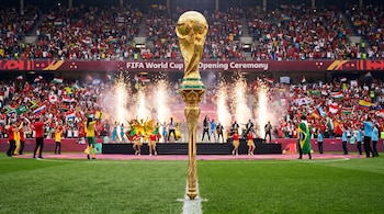 Trofeo de la Copa del Mundo FIFA en pedestal dorado en el campo. Detrás, ceremonia inaugural con bailarines, banda, pirotecnia y aficionados en gradas.