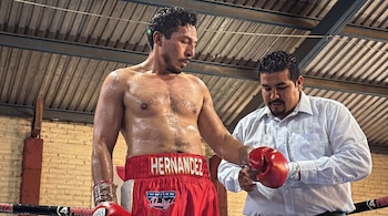 El boxeador Guillermo Hernández con pantalón rojo de boxeo junto a un hombre con camisa blanca que ajusta su guante rojo.