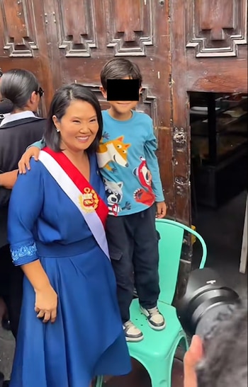 Keiko Fujimori con vestido azul y banda presidencial posa con un niño que viste camiseta azul con estampados y pantalón oscuro, frente a una puerta de madera