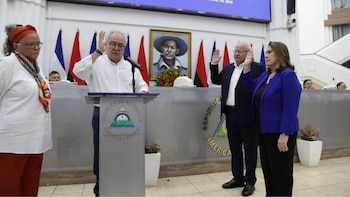 Un hombre habla desde un podio con el escudo de Nicaragua mientras una mujer en chaqueta azul y un hombre a su lado levantan la mano derecha