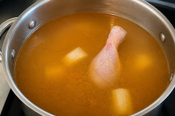 Una olla de acero inoxidable llena de caldo con una pierna de pollo y trozos de papa y yuca.