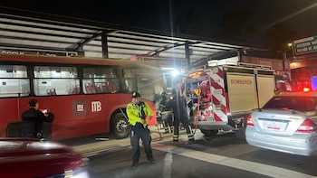 Un camión de bomberos y agentes atienden un autobús rojo detenido junto a una estación de transporte en una avenida por la noche