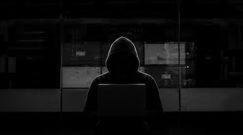 Silueta de una persona sentada con capucha usando una computadora portátil en un ambiente oscuro con patrones digitales borrosos en el fondo.