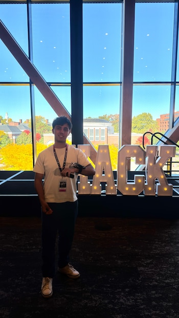 Un joven de pie junto a letras luminosas que forman la palabra HACK ante un gran ventanal que muestra árboles y edificios al exterior
