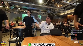 Un hombre en camisa blanca con gafas de sol en el cuello gesticula enérgicamente en un restaurante. Un hombre de negro lo observa de pie. Hay mesas y comensales al fondo